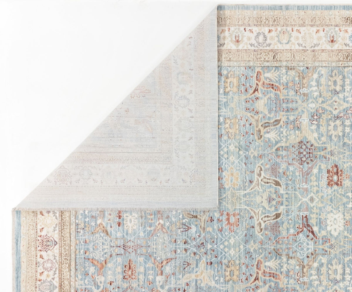 The Azure Oushak - Area Rug - APX2932 features a light blue Persian pattern with cream, beige, and brown accents, shown partially folded to display its lighter underside and intricate border designs.