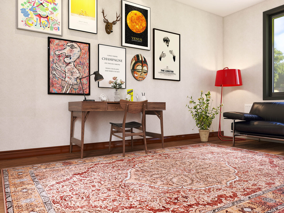 A home office includes a wooden desk, a chair, colorful artwork and framed posters on the wall, a red floor lamp, a plant, and the Baroque Charm - Washable Rug - JRN1016 (Custom Size) beneath a black sofa by a large window.
