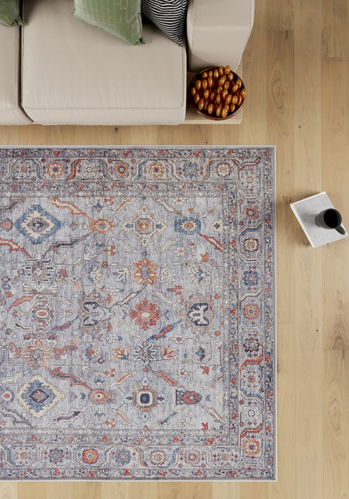 Load image 1 in gallery view. The Bellaris Gray Washable Rug - LCC3003 (Outlet) US, featuring blue, orange, and beige designs, lies on a wooden floor beside a beige sofa with green and black pillows. Nearby are a bowl of breadsticks and an open magazine with a cup.