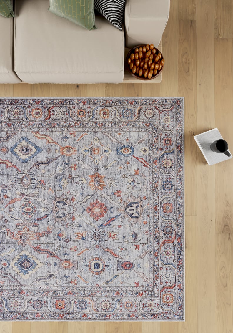 The Bellaris Gray Washable Rug - LCC3003 (Custom Size) lies on a light wood floor next to a beige sofa, a pillow, a bowl of pastries, and an open book with a cup of coffee.