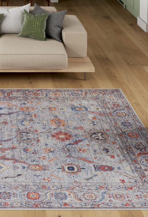 Load image 1 in gallery view. A modern living room with a light wood floor, a beige minimalist sofa with three green and black cushions, and a large Bellaris Gray Washable Rug - LCC3003 (Custom Size) in blue, red, and beige tones.