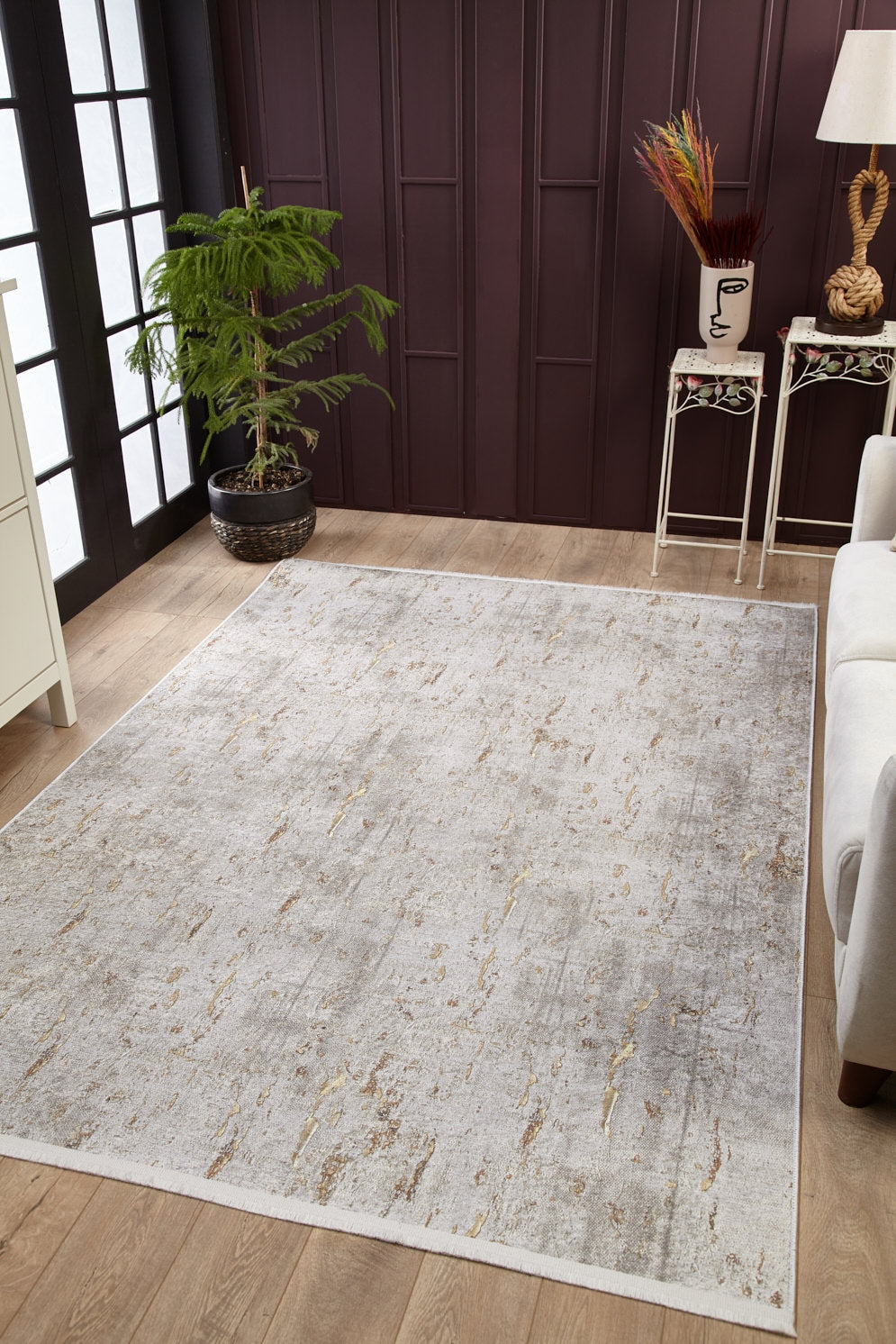 A modern living room features the Berry Blossom - Washable Rug - JR1818 (Custom Size) on wooden floors, a white sofa, a potted plant by the window, and a side table with decor and a face-shaped vase against a dark paneled wall.