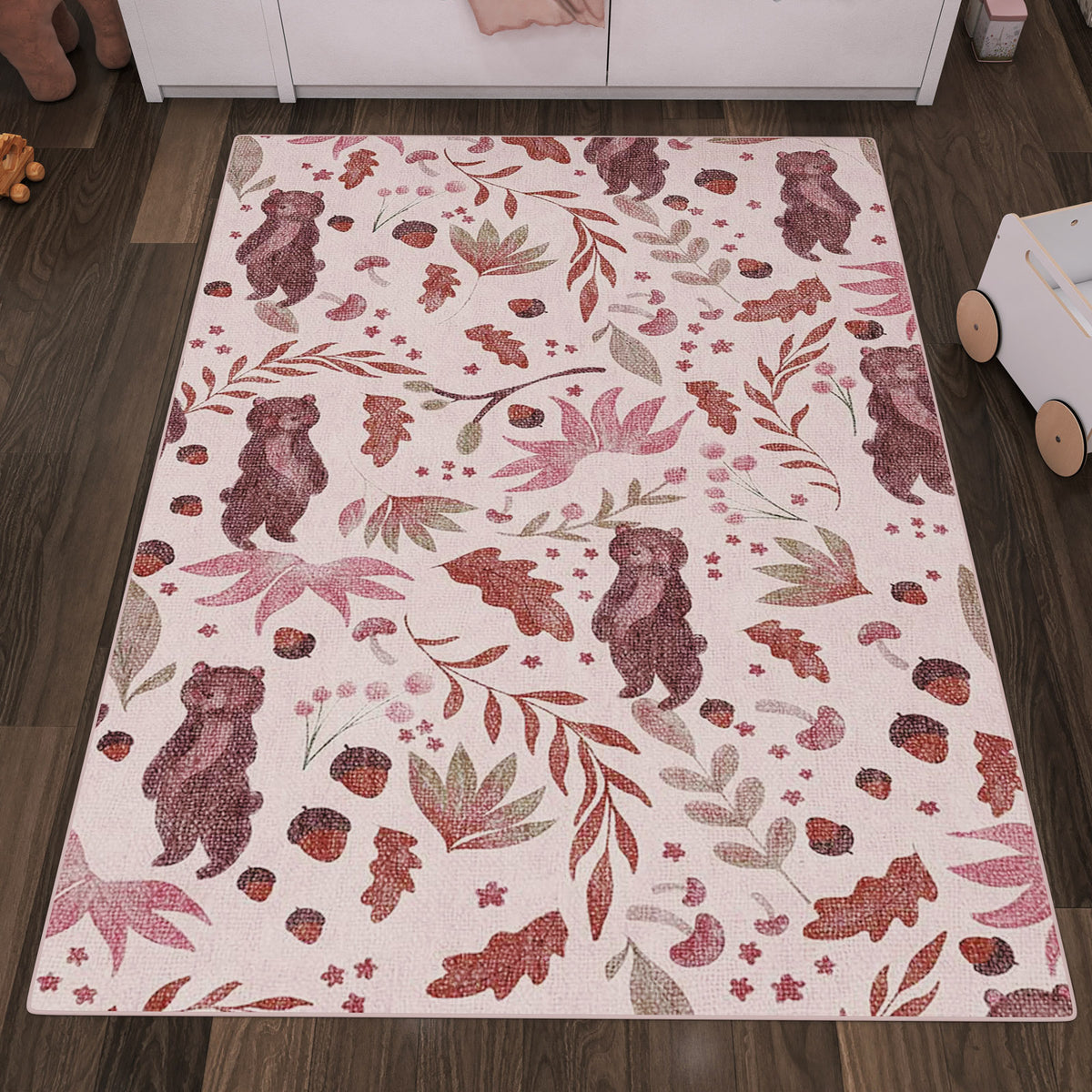 Pink rug with a pattern of brown bears, acorns, and leaves, styled in a nursery with dark wood flooring.