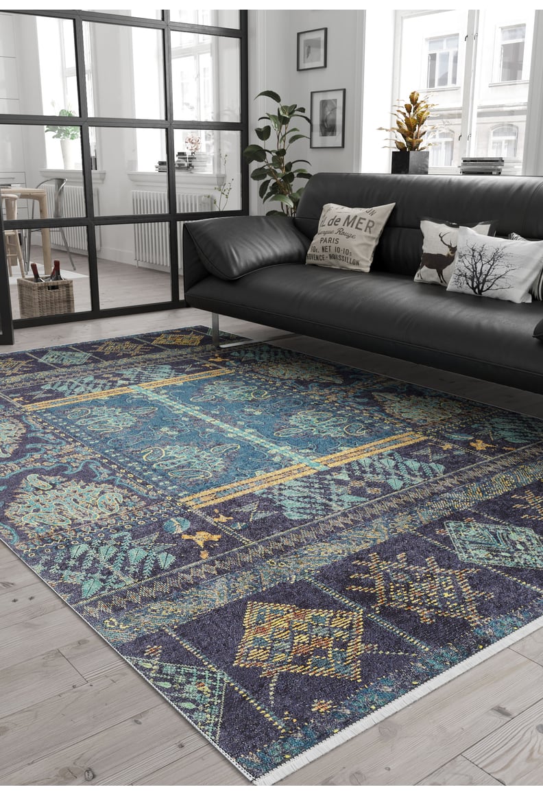 A modern living room with a black leather sofa, decorative pillows, the Blossoming Bazaar - Washable Rug - JR1862 in blue for high-traffic areas, potted plants, natural light from large windows, and black-framed glass partitions dividing the space.