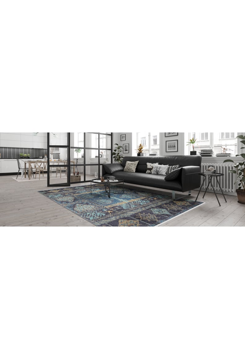 A modern living room features a black sofa, decorative pillows, and the Blossoming Bazaar - Washable Rug - JR1862 in blue—perfect for high-traffic areas. Large windows fill the space with light; plants near the radiator and an open dining area add charm.