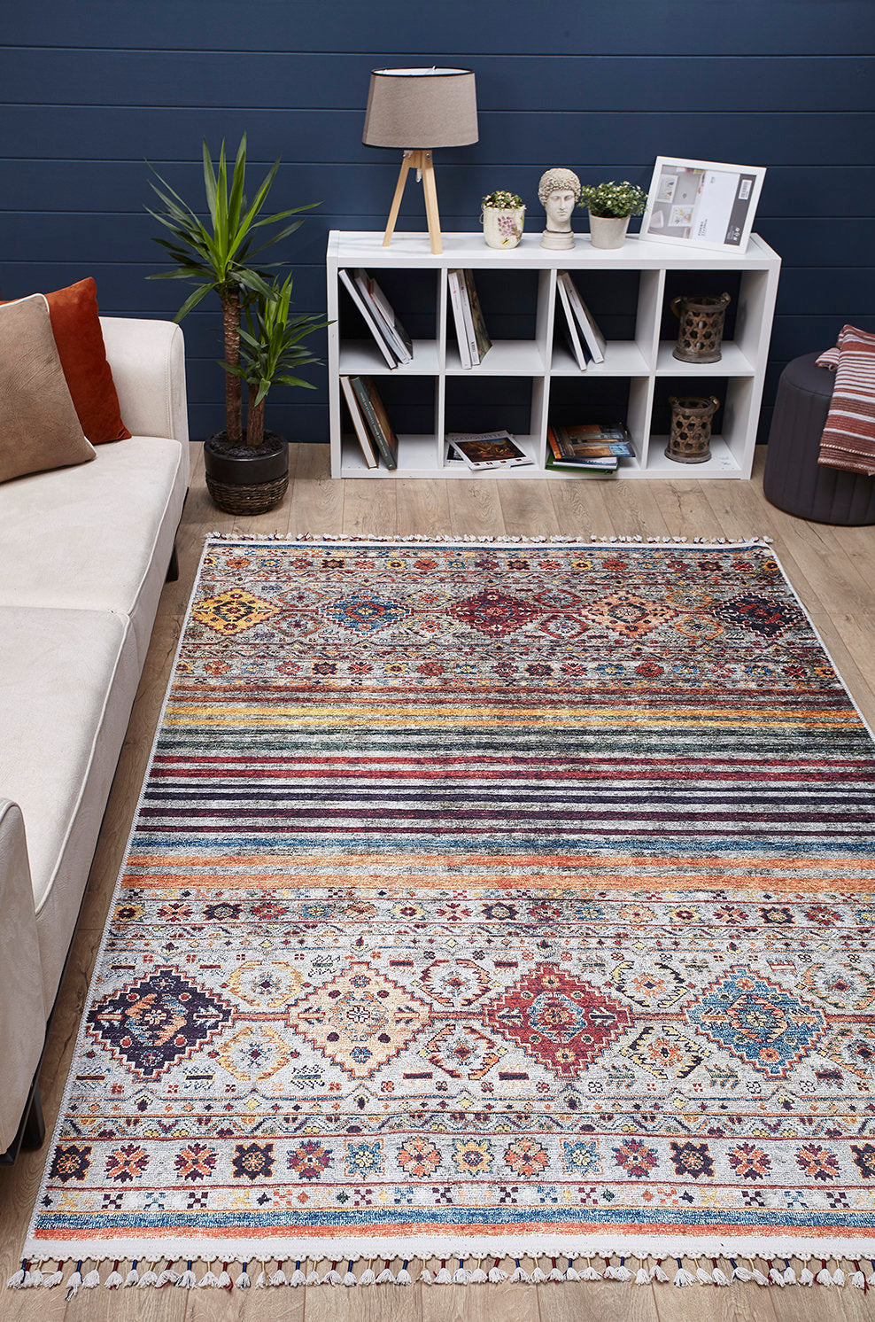 A modern living room features the Bohemian Bouquet - Washable Rug - JR5090 (Custom Size), a beige sofa with cushions, potted plant, white cube bookshelf with decor and books, and a table lamp set against a navy blue wall.