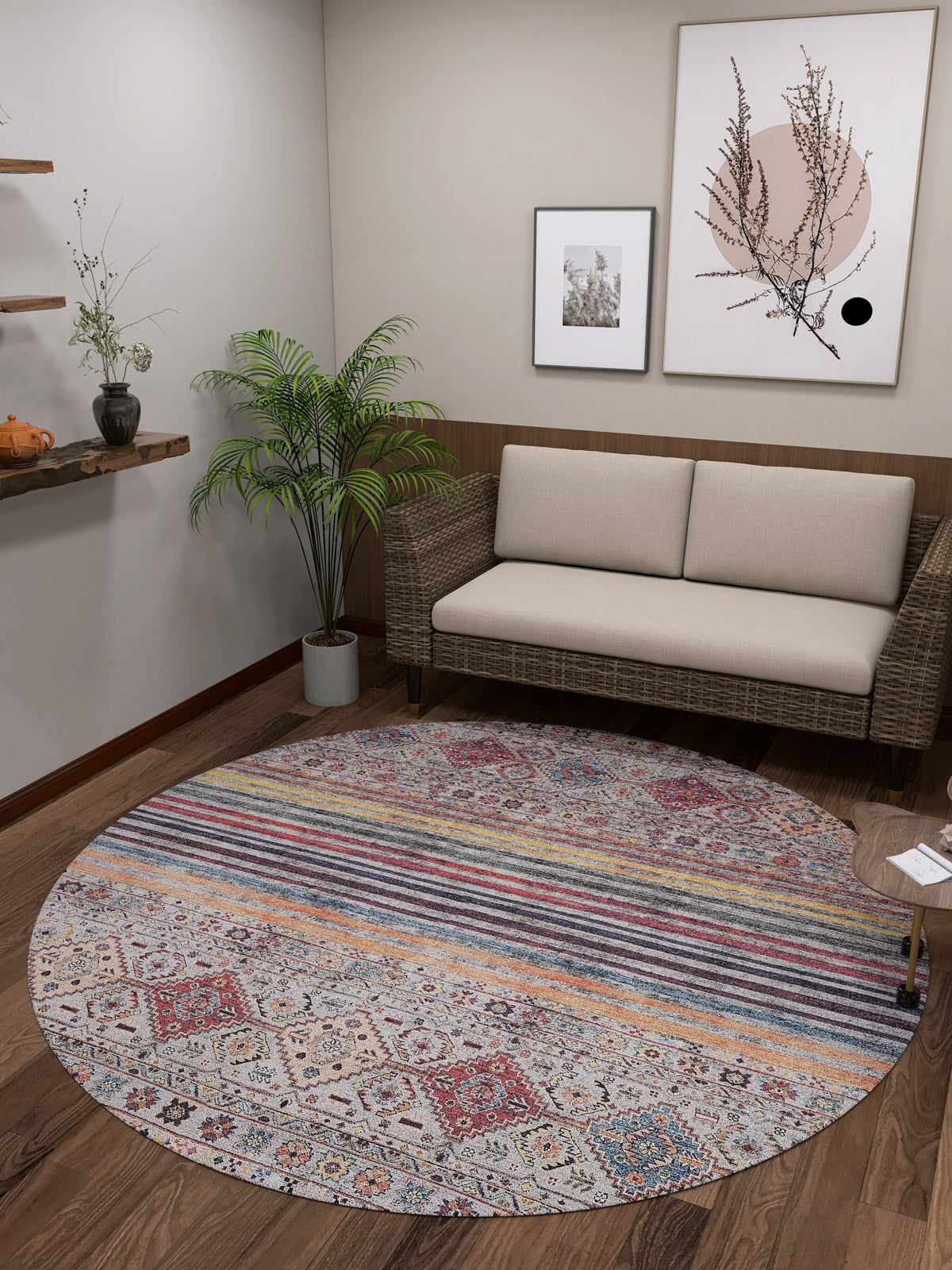 A cozy living room features the Bohemian Bouquet - Washable Rug - JR5090 (Custom Size), a beige wicker-frame sofa, a potted plant, wooden shelves with decor, minimalist framed art, and wood flooring.