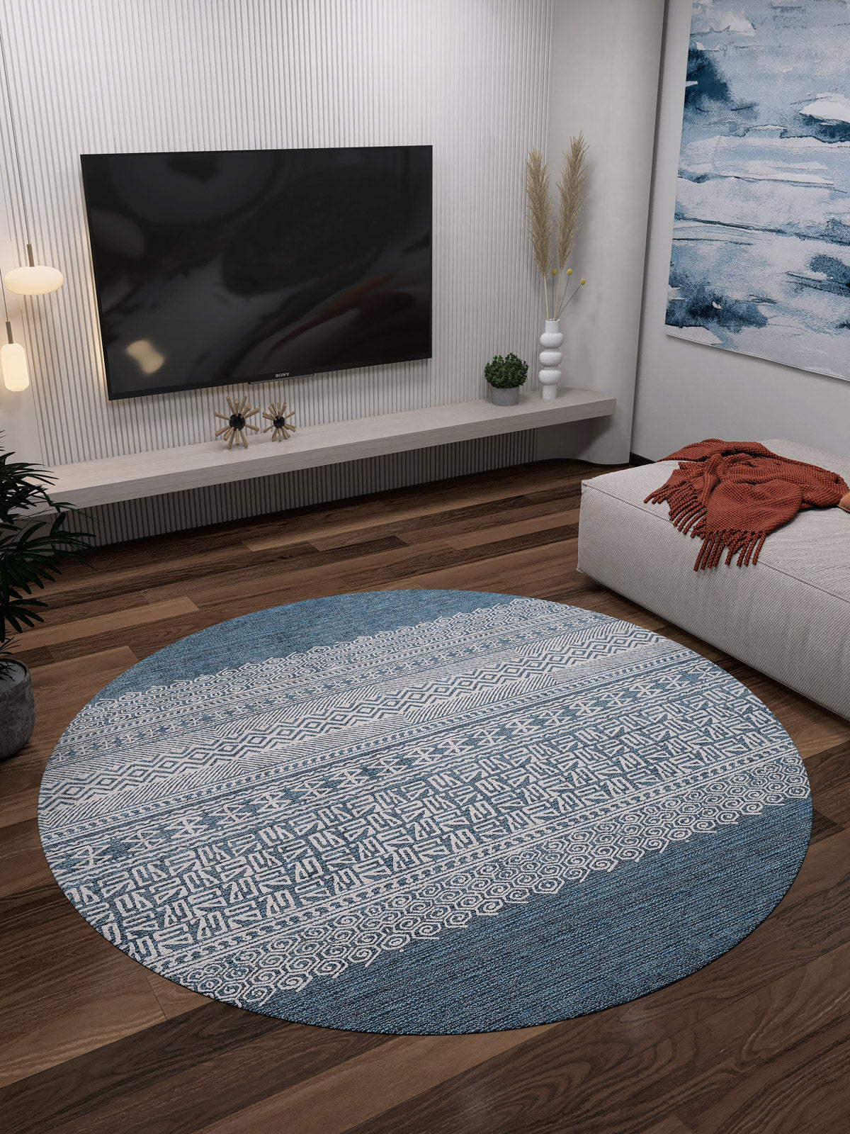A modern living room features the Bohemian Retreat - Washable Rug - JR1589 (Custom Size), round in blue and white, ideal for high-traffic areas, on a dark wood floor with a wall-mounted TV, low decor shelf, abstract painting, and beige ottoman with an orange blanket.