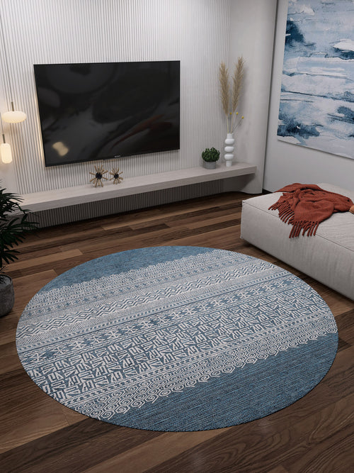Load image 1 in gallery view. A modern living room features the Bohemian Retreat - Washable Rug - JR1589 (Custom Size), round in blue and white, ideal for high-traffic areas, on a dark wood floor with a wall-mounted TV, low decor shelf, abstract painting, and beige ottoman with an orange blanket.