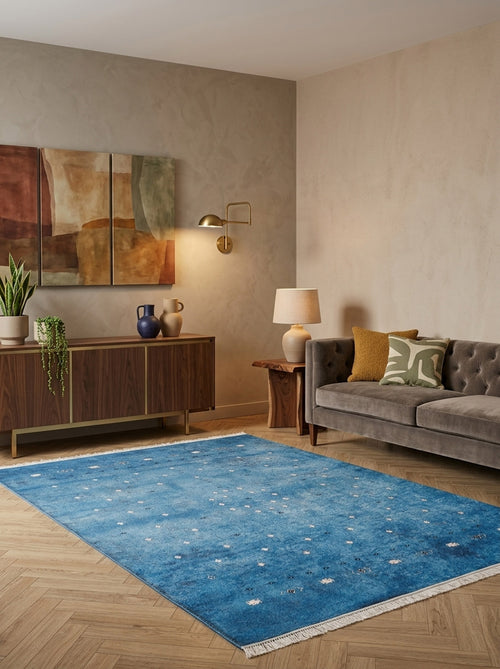 Load image 1 in gallery view. A modern living room with the Bohemian Whirlwind - Washable Rug - JR1989 in blue for high-traffic areas, a gray tufted sofa with patterned cushions, a wooden sideboard with plants and vases, abstract wall art, and wooden tables with lamps.