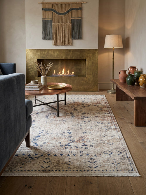 Load image 1 in gallery view. Distressed beige rug with blue and rust-colored traditional patterns, styled in a living room with a brass fireplace.