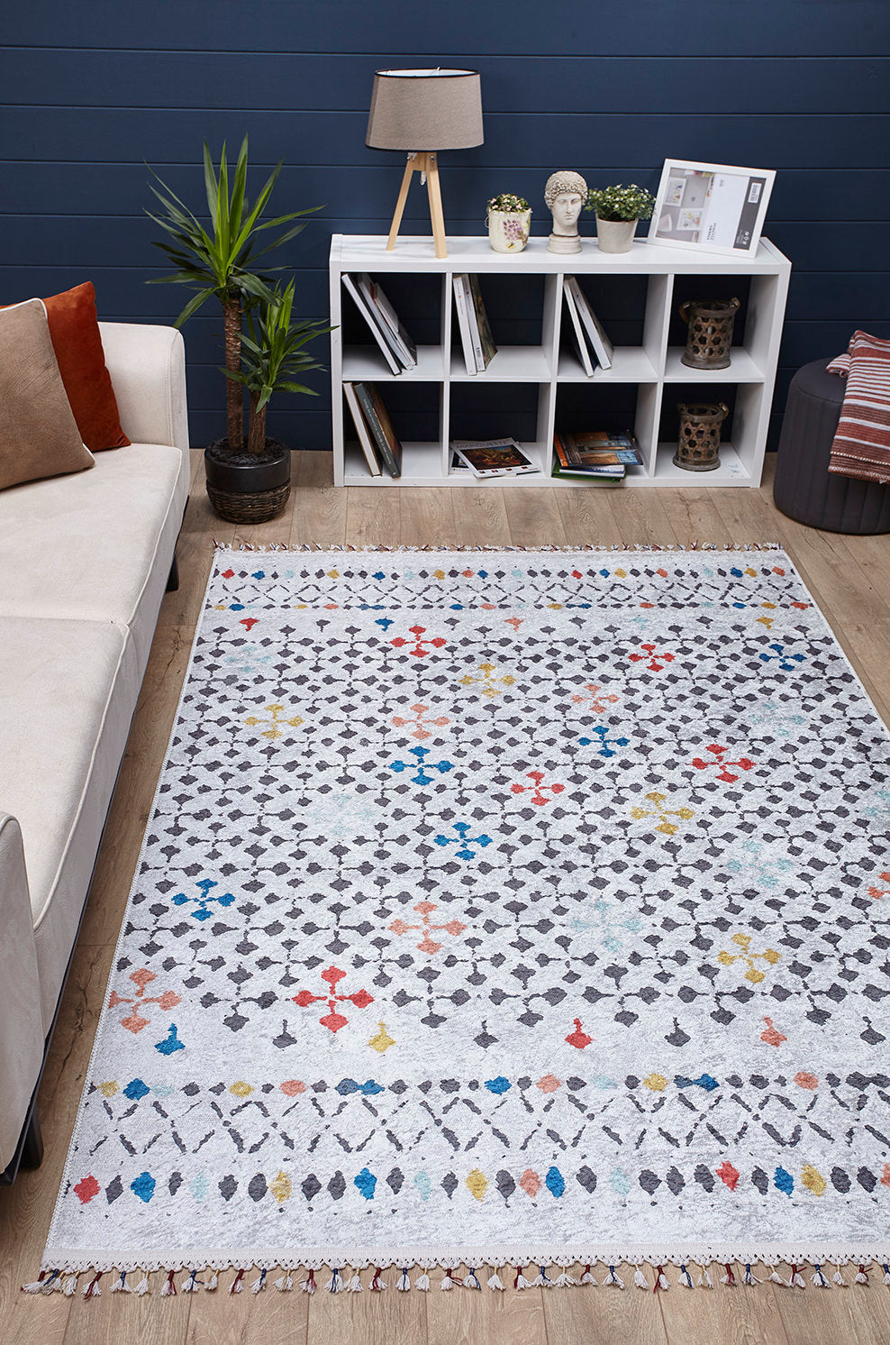 A modern living room featuring the Boho Breeze - Machine-Washable Rug - JR5011 (Custom Size) with geometric, colorful accents, a beige sofa with cushions, a potted plant, and a white cubed bookshelf against a dark blue wall.