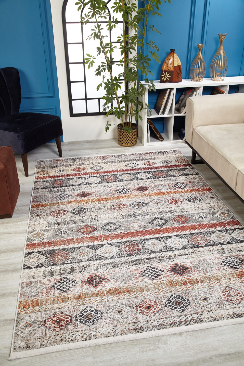 A modern living room featuring the Boho Oasis - Washable Rug - JR1618 (Custom Size) in gray-anthracite earth tones, paired with a beige sofa, black chair, tall green plant, vases on shelves, and a blue accent wall—perfect for busy spaces.