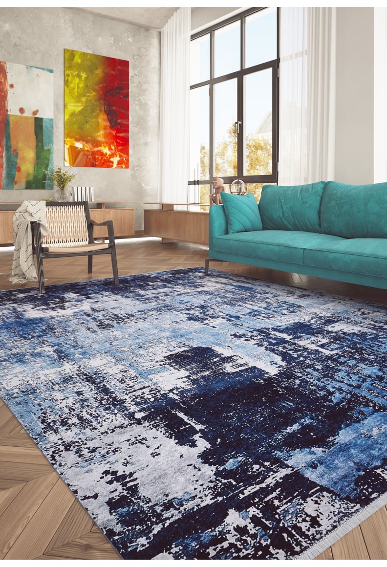 A modern living room features the Bold Blooms - Washable Rug - JR836 in blue and white, a teal sofa, wooden chair, and large windows. Abstract art on a concrete wall creates a cozy, vibrant space ideal for high-traffic areas.