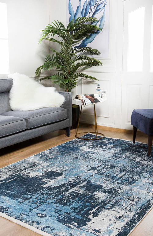 Load image 1 in gallery view. A modern living room with the Bold Blooms - Washable Rug - JR836 (Custom Size) in blue, featuring a gray sofa with a white faux fur throw, potted plant, small decor side table, blue upholstered stool, and wooden flooring.