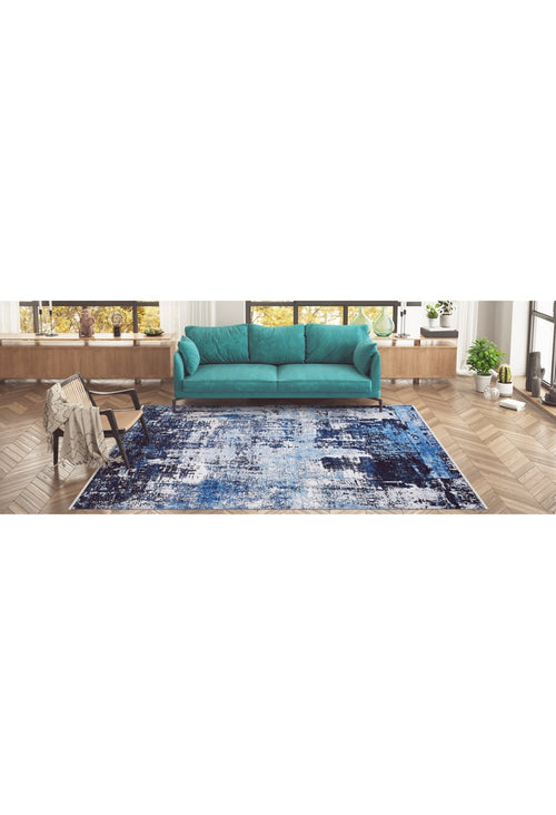 Load image 1 in gallery view. A modern living room features a teal sofa, wooden floors, large windows, and plants on a low shelf. The Bold Blooms - Washable Rug - JR836 (Custom Size) in blue adds style—perfect for high-traffic areas. A beige throw and books rest on a side chair.