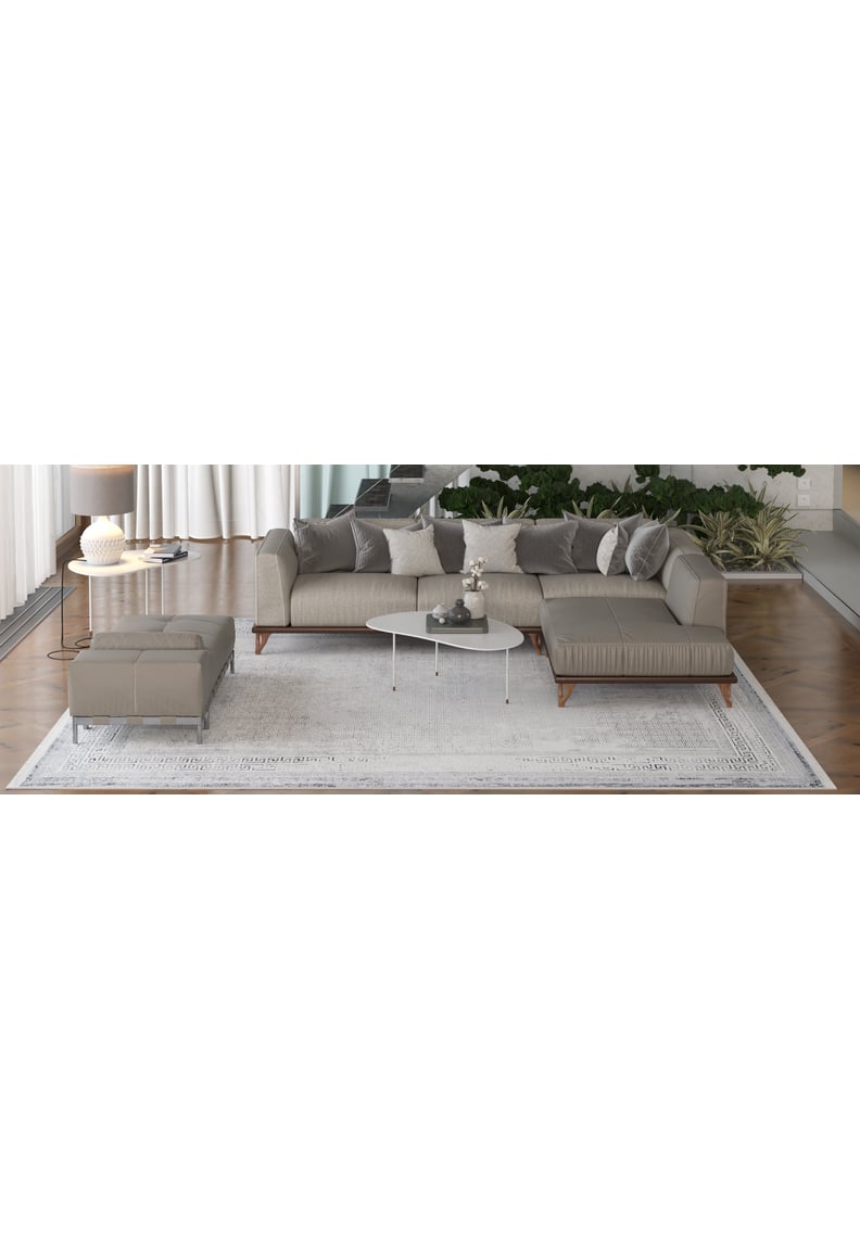 A modern living room features a gray sectional, ottoman, white oval coffee table, décor items, and the Bold Borderline - Washable Rug - JR1984—ideal for high-traffic areas. Green plants and sheer curtains brighten the space.