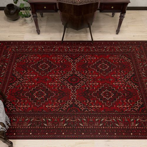 Load image 1 in gallery view. The Bold Thread - Kilim - BHR05 is a rectangular red Turkish rug with geometric and floral patterns, made of soft chenille-polyester fibers. It’s shown on a light wood floor by a dark wooden desk and chair, with a plant nearby.