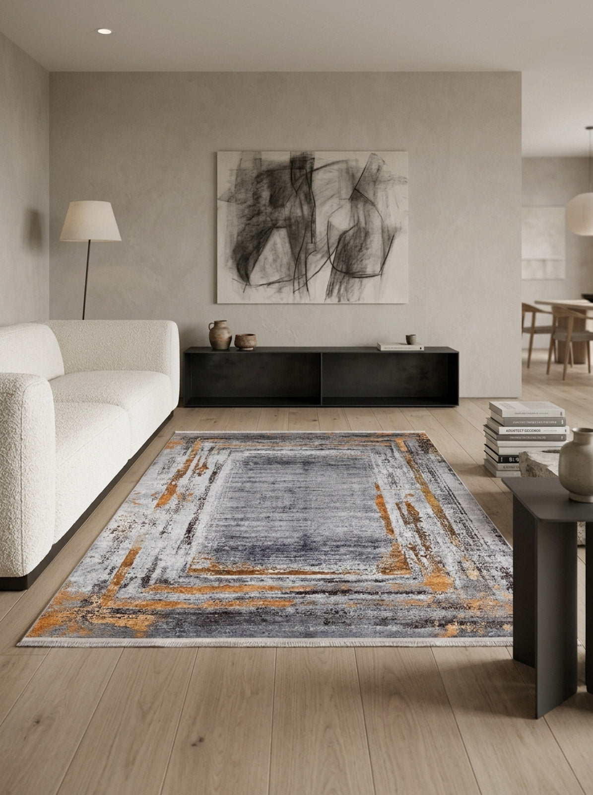 A modern living room features light wood flooring, a textured white sofa, abstract wall art, a black low console, floor lamp, and the Bold and Chic - Washable Rug - JR1198 in gray, orange, and cream—ideal for high-traffic spaces. Dining area in background.