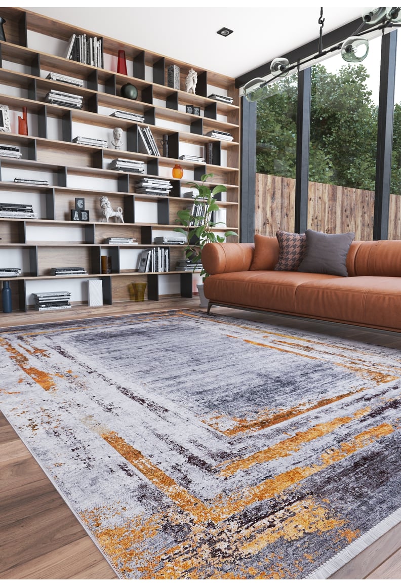 A modern living room with a large window, leather sofa, and the Bold and Chic - Washable Rug - JR1198 in gray, orange, and white—an allergy-friendly choice. Floor-to-ceiling shelves hold books and decor; trees are visible outside.