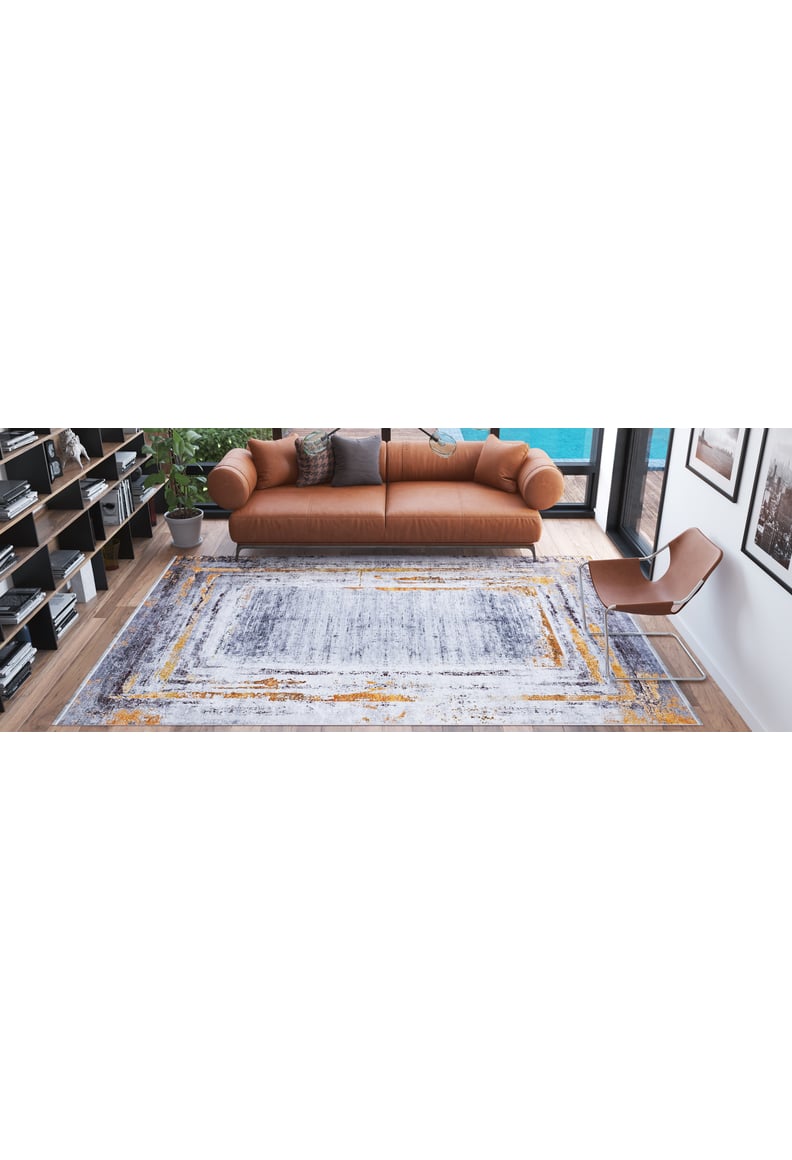 A modern living room featuring a brown leather sofa, gray and brown cushions, the Bold and Chic - Washable Rug - JR1198 (Outlet) US with gray and yellow patterns, a potted plant, a brown accent chair, and bookshelves along one wall.