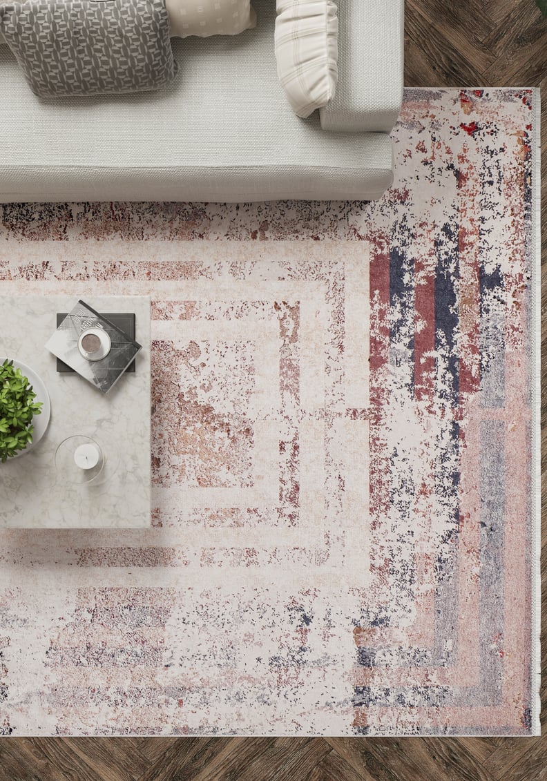 A modern living room viewed from above shows a light gray couch, marble coffee table with a plant, drink, and magazines, on the JR1410 Bold and Geometric washable rug—ideal for high-traffic areas—laid over herringbone wood floors.