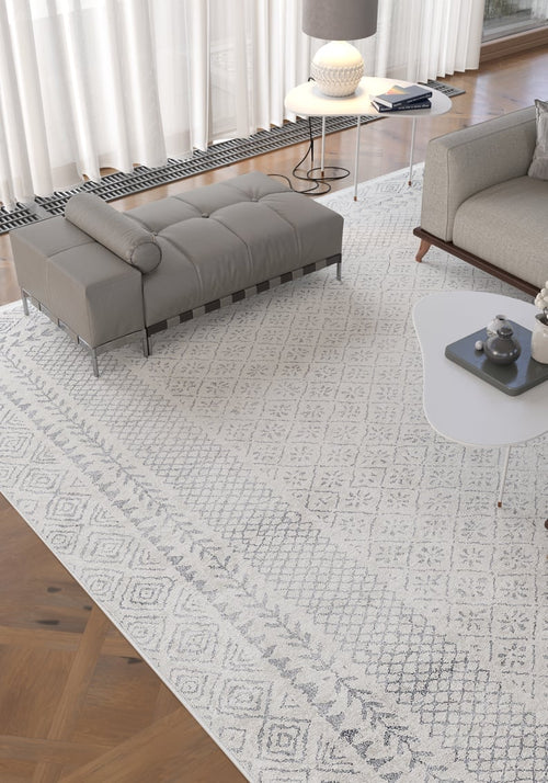 Load image 1 in gallery view. A modern living room features the Borderline Brilliance - Washable Rug - JR1987, ideal for high-traffic areas, paired with a gray chaise lounge, white side table with lamp and book, light gray sofa, and sheer curtains letting in natural light.