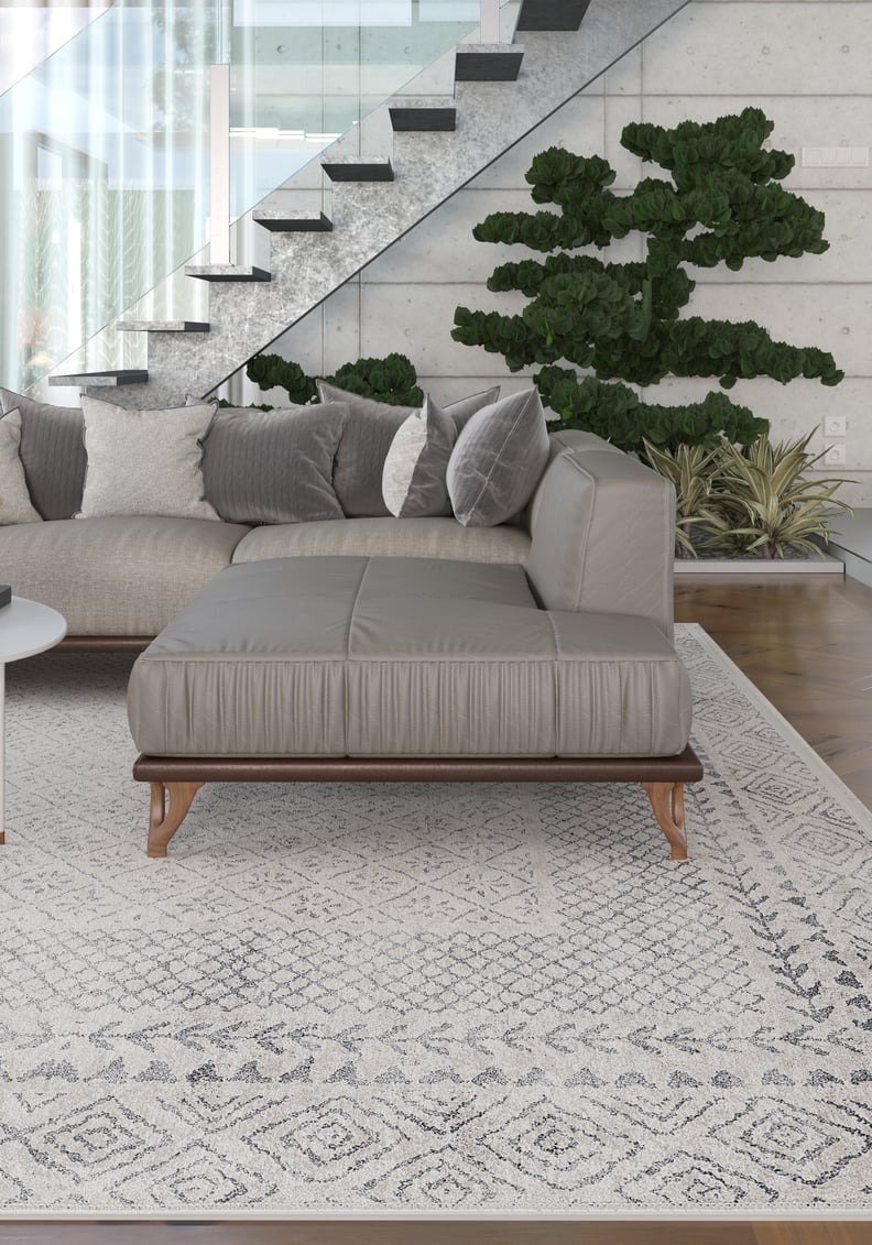 A modern living room with a gray sectional, decorative pillows, and potted plants features the Borderline Brilliance - Washable Rug - JR1987 (Custom Size) in gray-anthracite—ideal for high-traffic areas. A glass-railed staircase is in the background.
