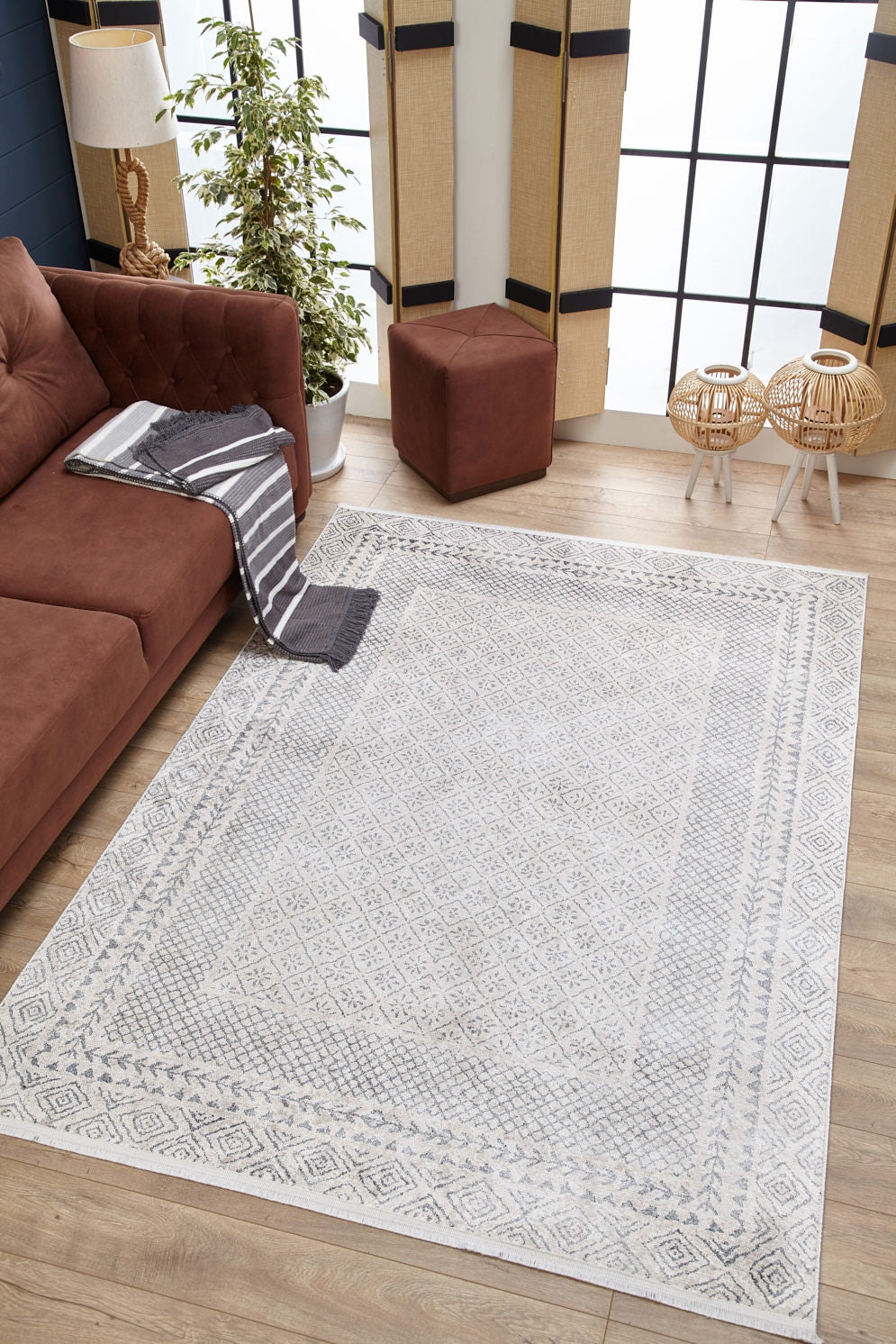 The Borderline Brilliance - Washable Rug - JR1987 (Custom Size) in gray-anthracite is on a wooden floor beside a brown sofa with striped blanket, near a potted plant, ottoman, and wicker tables—perfect for high-traffic spaces.