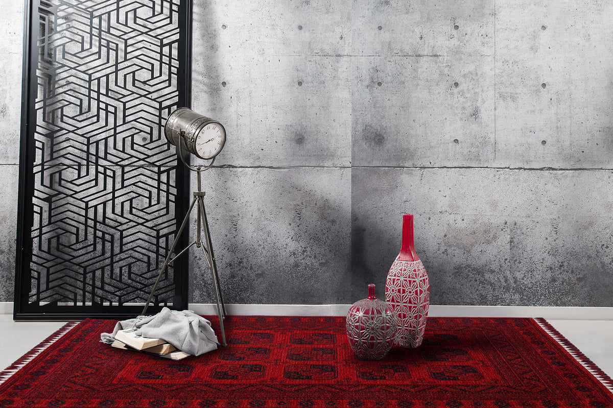 A modern interior featuring a gray concrete wall, a black geometric panel, a vintage floor lamp, two patterned vases with red accents, the Cherry Texture - Washable Kilim - BHR01 in red patterns, and a stack of books with gray cloth.