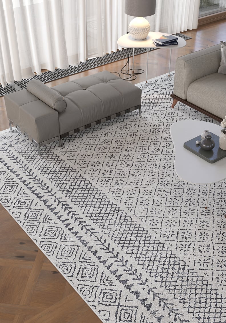 A modern living room features the Classic Contrast - Washable Rug - JR1985 in gray-anthracite, a gray bench and sofa, a white coffee table, side table with lamp, and large windows with sheer white curtains.