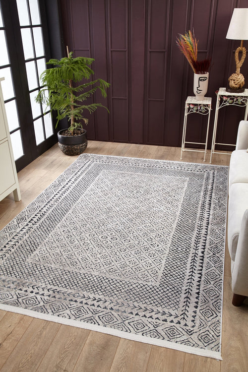 Load image 1 in gallery view. A living room with wooden floors features the Classic Contrast - Washable Rug - JR1985 (Custom Size) in gray-anthracite, a potted plant by large windows, a white sofa, and decorative tables with stylish accents against a dark paneled wall.