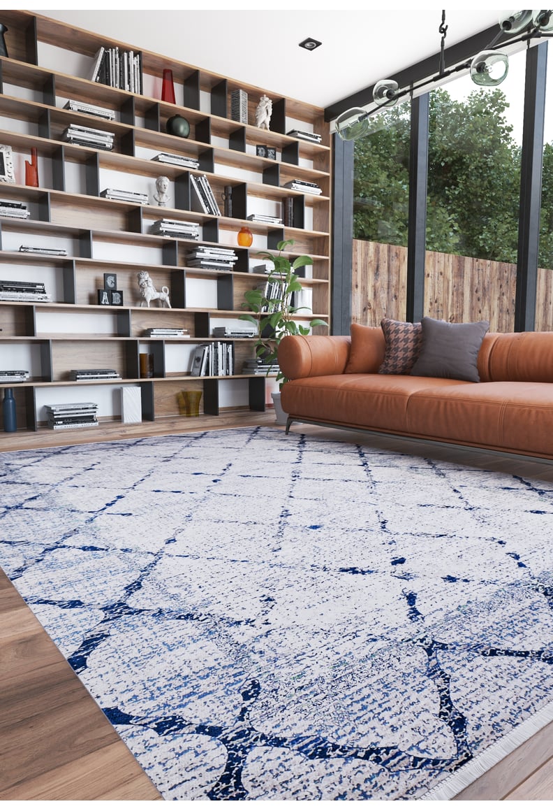 A modern living room with a tan leather sofa, gray pillows, the Classic Trellis Washable Rug (JR710) in blue and white, floor-to-ceiling shelves filled with books and decor, and tall windows overlooking greenery.