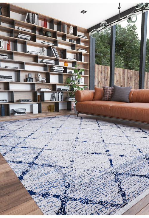 Load image 1 in gallery view. A modern living room with a tan leather sofa, gray pillows, the Classic Trellis Washable Rug (JR710) in blue and white, floor-to-ceiling shelves filled with books and decor, and tall windows overlooking greenery.