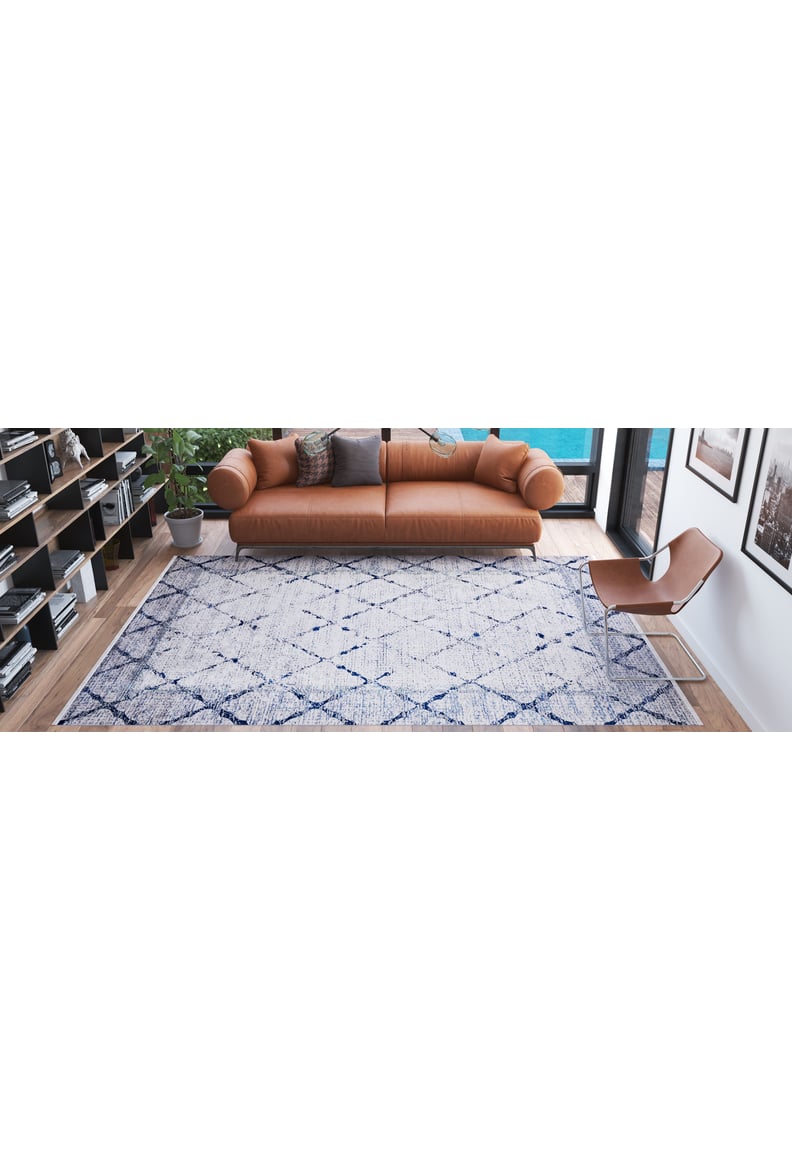 A modern living room with a tan leather sofa, brown chair, Classic Trellis - Washable Rug - JR710 in blue and white, bookshelves, a potted plant, and large windows letting in natural light.