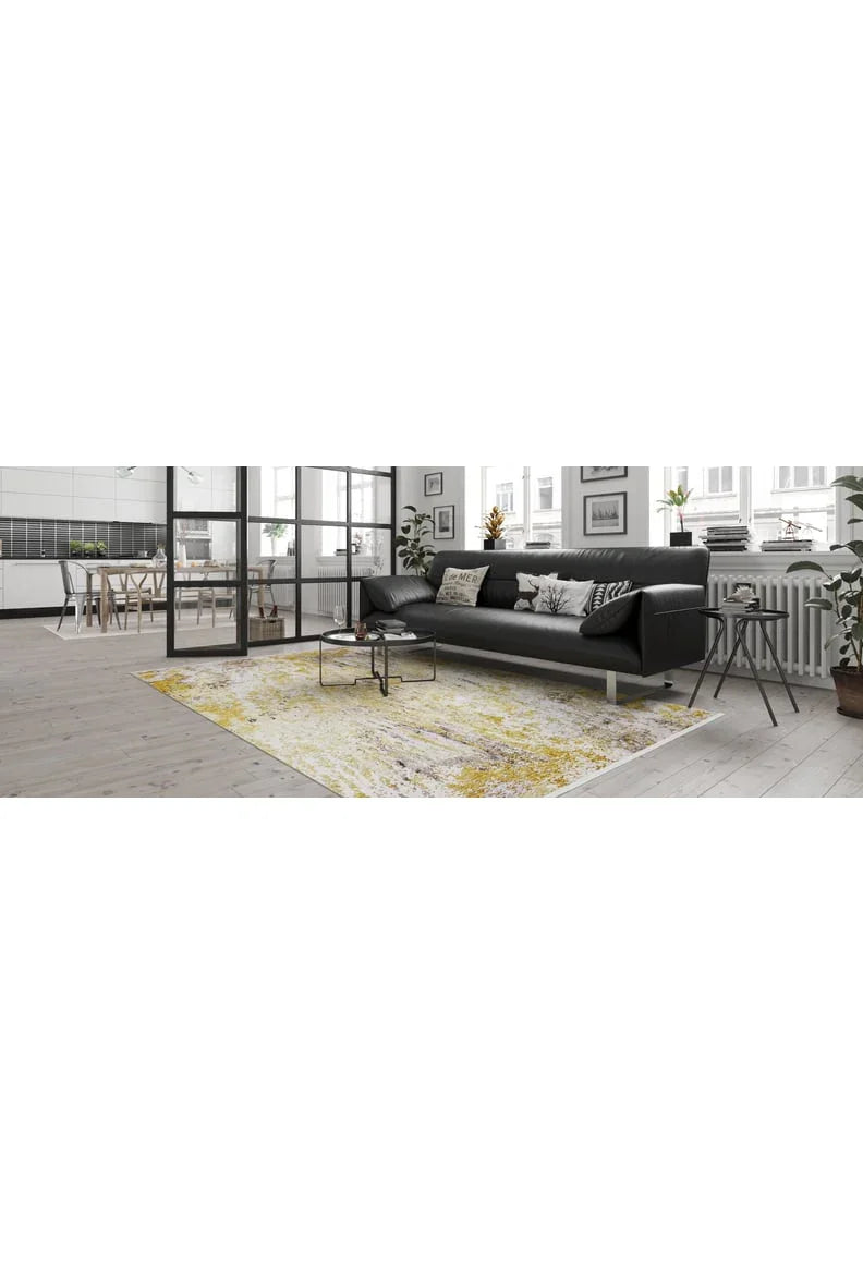 A modern living room with a black sofa, decorative cushions, and the Color Splash - Washable Rug - JR146 in yellow-gold on light wood floors. Large windows fill the space with natural light, and an open dining area and kitchen enhance its high-traffic appeal.