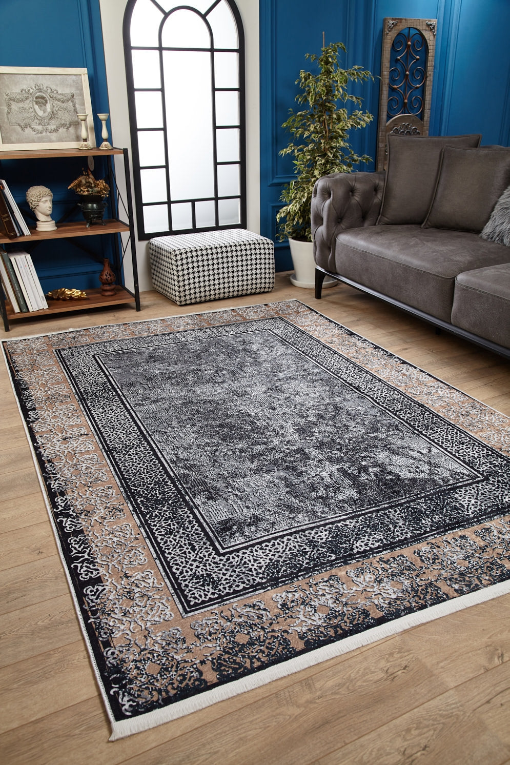 A modern living room features the Contemporary Borderline - Washable Rug - JR1798 (Custom Size) in gray and beige on wooden floors, ideal for high-traffic areas, paired with a gray sofa, black-and-white ottoman, plant, bookshelf, and tall arched window.