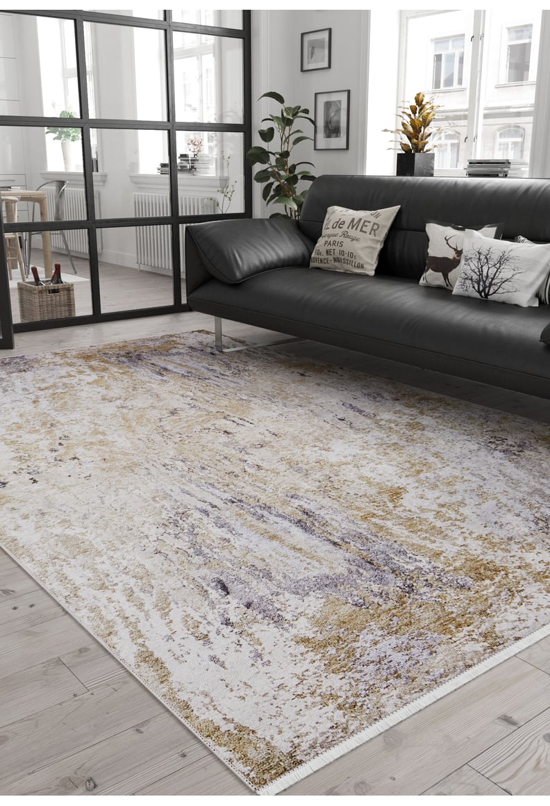 A modern living room with a black leather sofa, pillows, a potted plant, and the Contemporary Canvas - Washable Rug - JR145 (Custom Size) in neutral tones on light wood floors. Large windows fill the space with natural light—perfect for busy areas.