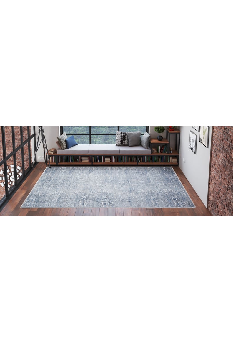 A spacious, modern room with wooden floors and exposed brick walls features the Contemporary Serenity - Machine-Washable Rug JR1884 (Custom Size) in blue—ideal for high-traffic areas—plus a bench with cushions and books by large windows.