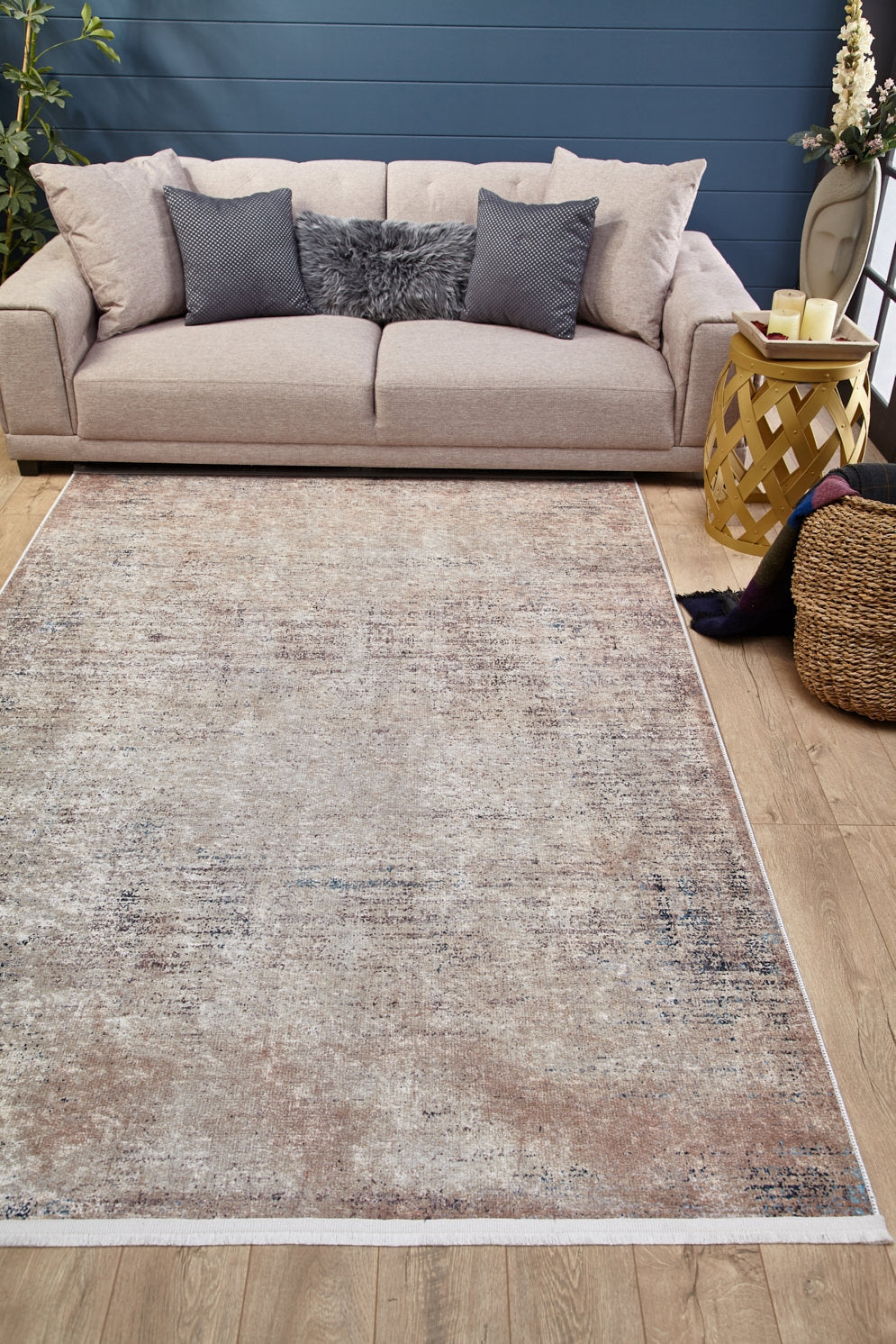 A beige sofa with gray and fur pillows sits against a blue wall. In front, the Copper Glow - Washable Rug - JR1700 (Custom Size), ideal for high-traffic areas, rests on the wood floor beside a woven basket and yellow side table with candles and flowers.