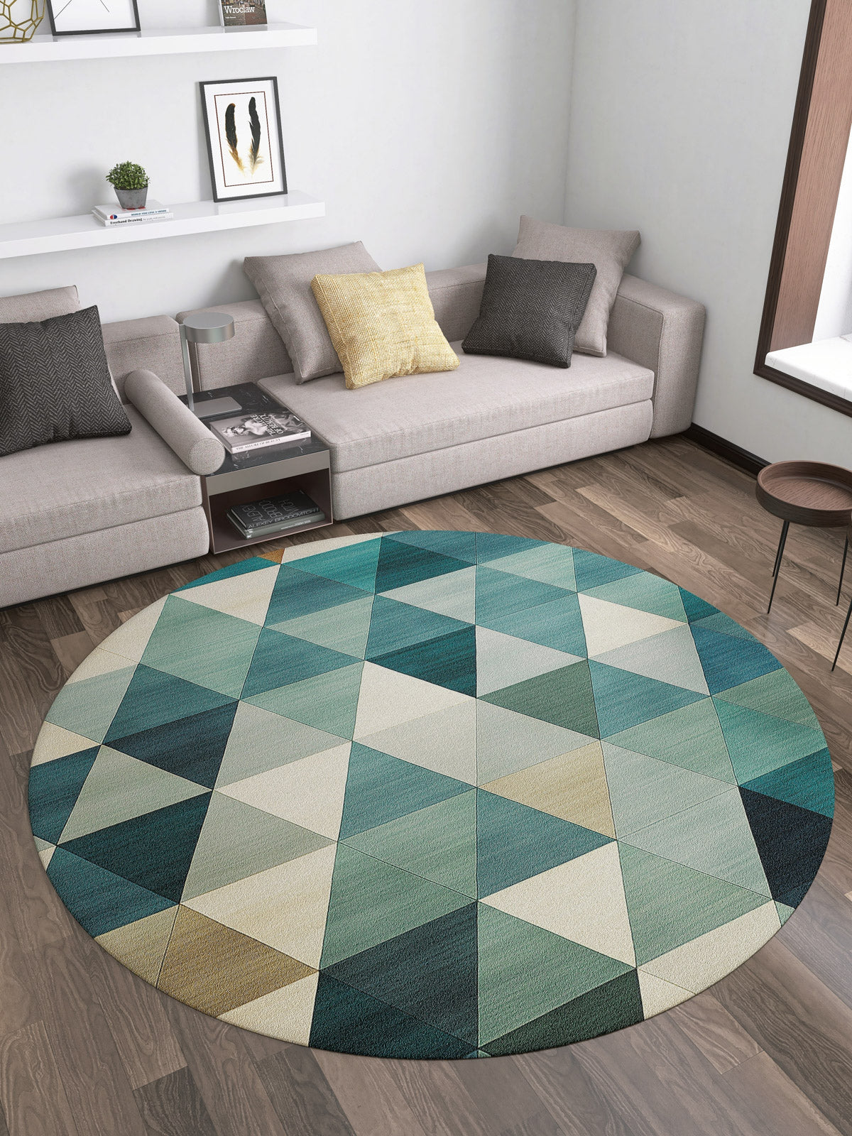 A modern living room with a gray sectional and neutral cushions features the Cubism Collage Blue Washable Rug - MDJ008 (Custom Size), showcasing teal, green, and beige geometric patterns—ideal for high-traffic spaces—on a wooden floor. Books and art line wall shelves above.