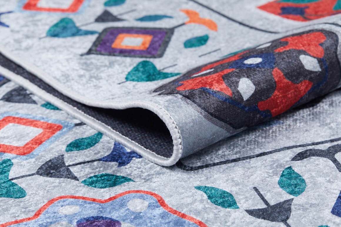 A close-up of the Desert Drift - Washable Rug - JR1523 (Custom Size) shows its vibrant abstract geometric and floral patterns on a light gray background. The folded corner reveals detailed texture and stitching, ideal for high-traffic areas.