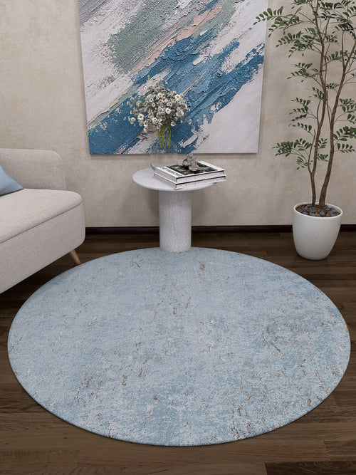 Load image 1 in gallery view. A modern living room features the Earthy Sands - Washable Rug - JR1819, perfect for high-traffic areas, a small white side table with books and flowers, a potted plant, a beige sofa, and an abstract blue and white painting on the wall.