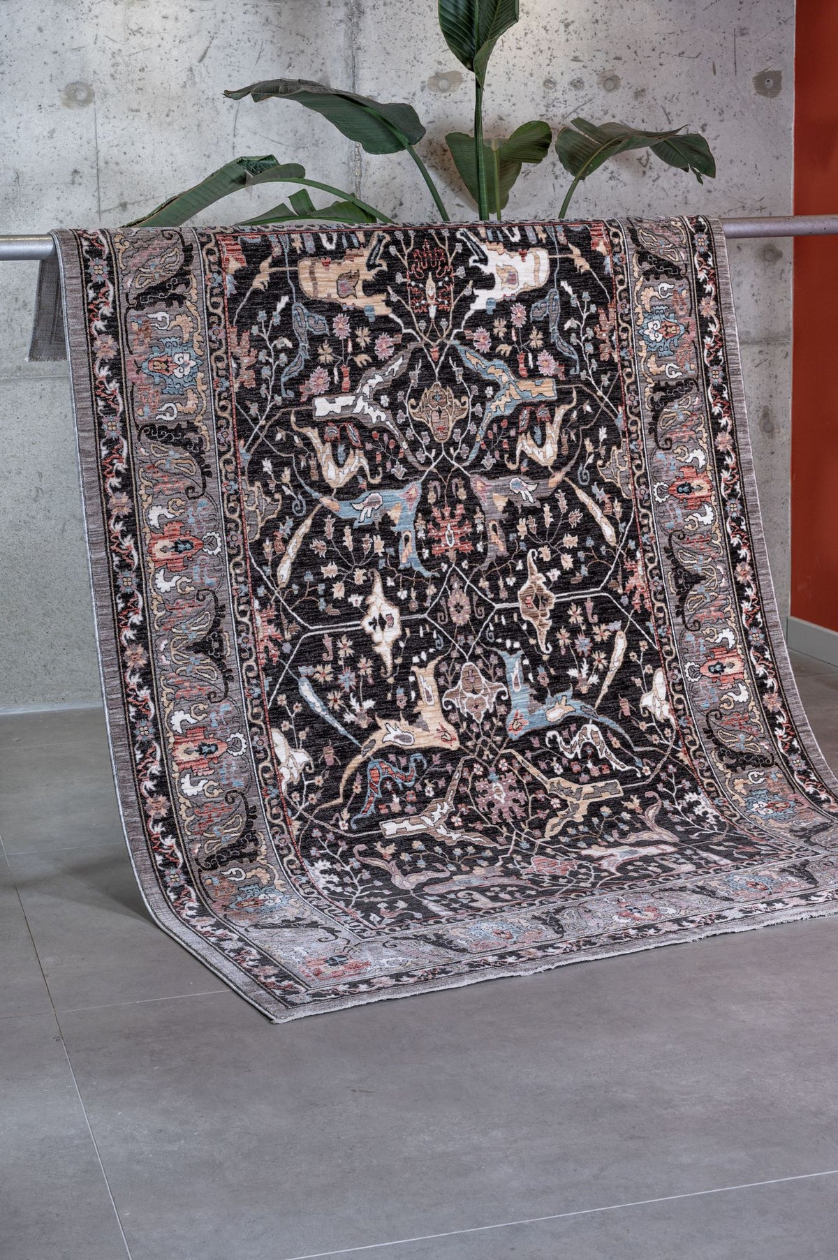 Black traditional rug with intricate floral and animal motifs in muted blue, pink, and cream, draped in a studio setting.