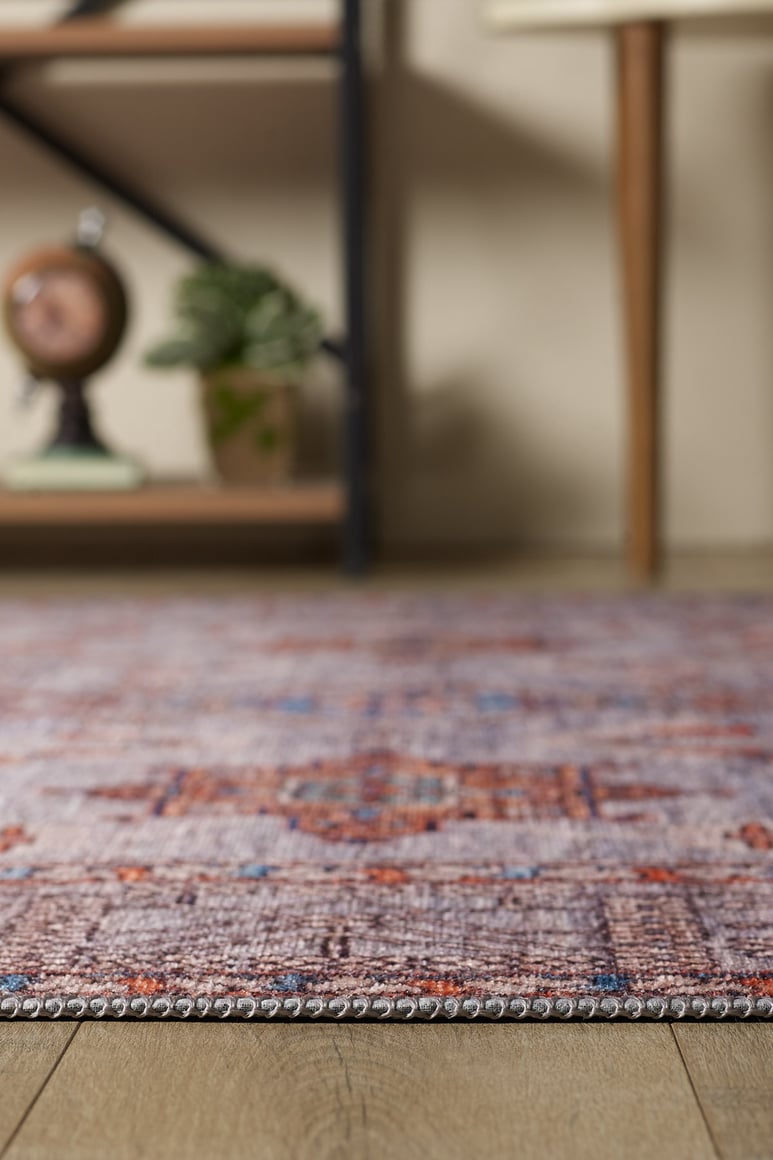 A close-up of the Echelon Bordered Washable Rug - LCC3018, featuring vibrant red, purple, and orange patterns on a wooden floor; a plant, clock, and wooden furniture appear blurred in the background—ideal for high-traffic areas.