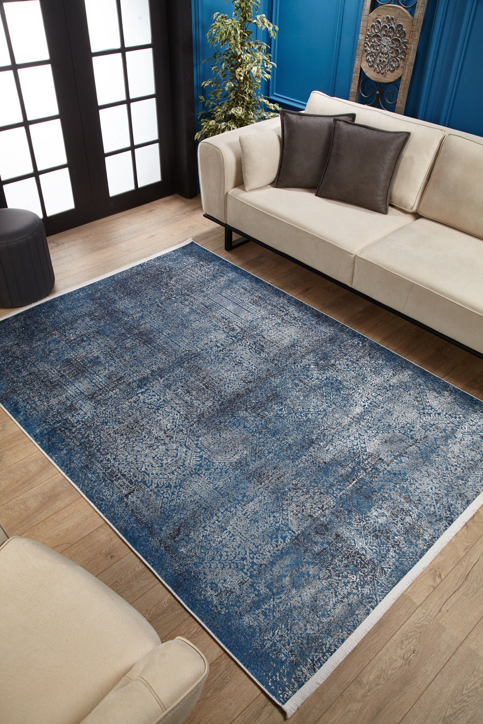 A modern living room with beige sofas, dark pillows, and the Elegant Vine - Washable Rug - JR1847 (Custom Size) in blue for high-traffic wooden floors, plus a corner plant, blue walls, and black-framed glass doors.