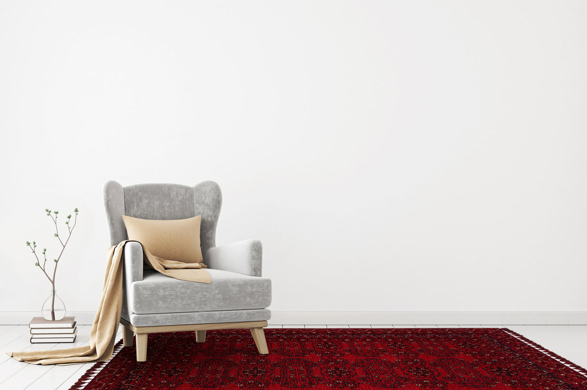 A gray armchair with a beige pillow and throw sits on the Ember Stitch - Washable Kilim - BHR02 rug in a minimalistic room. A small stack of books and a plant are placed on the floor beside the chair.