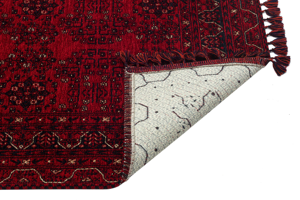 The Ember Stitch - Washable Kilim - BHR02 is a red patterned Turkish rug with fringed edges and a geometric border. One corner is flipped, showing the light-colored underside with a distinct pattern. The background is white.