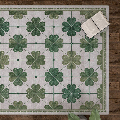 Load image 1 in gallery view. The Emerald Charm - Washable Rug - JRS013, featuring green four-leaf clovers and a subtle floral pattern, lies on a wooden floor with books and plant leaves nearby—ideal for high-traffic areas.