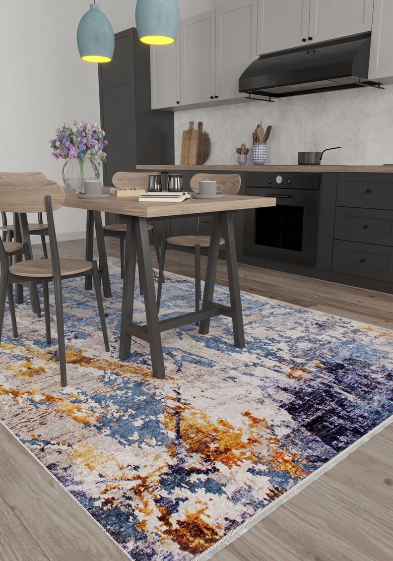 A modern kitchen and dining area with light wood furniture, gray cabinets, and a stovetop. The Enigmatic Elegance - Washable Rug - JR787 (Custom Size) in vibrant blue, orange, and yellow covers the floor—ideal for high-traffic areas. A vase with purple flowers sits on the table.